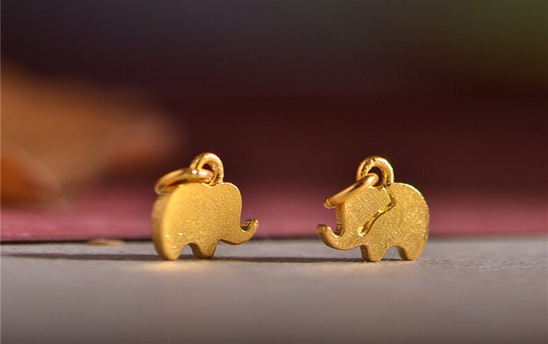 May include: Two small, gold-colored elephant charms with textured surfaces. Each charm has a loop at the top for attaching to a chain or earring hook. The elephant charms are displayed against a blurred, colorful background.