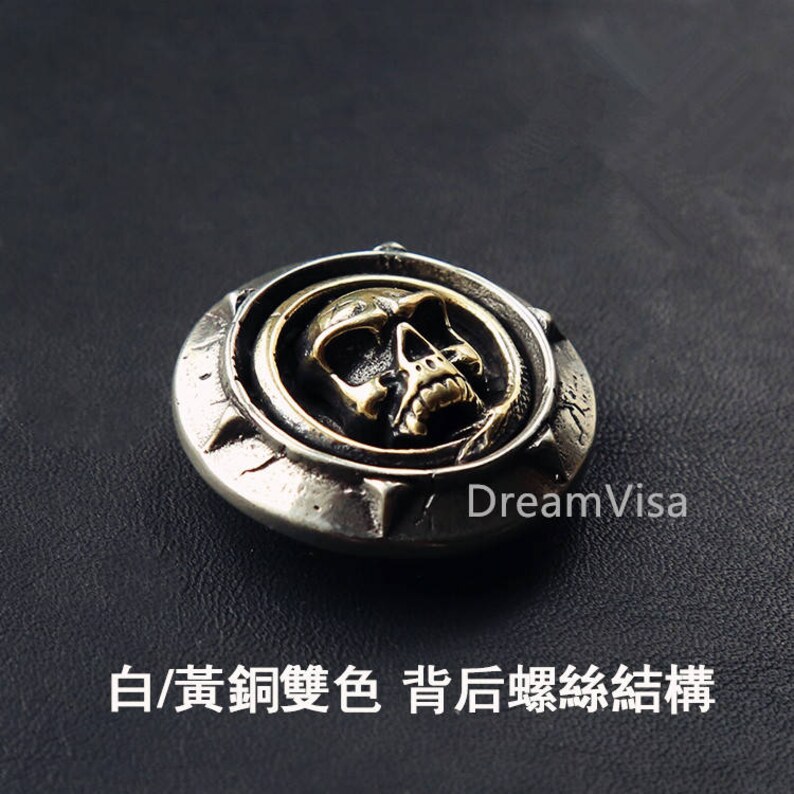May include: A silver and gold skull button with a detailed design. The skull is in the center of the button and is surrounded by a circle of silver.