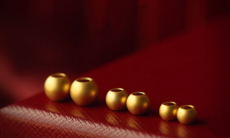 K&ouml;nnte beinhalten: Eine Reihe von sechs goldfarbenen Metallperlen in verschiedenen Gr&ouml;&szlig;en, die von der gr&ouml;&szlig;ten bis zur kleinsten auf einer roten Oberfl&auml;che angeordnet sind.