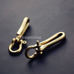 Op de afbeelding: Twee goudkleurige metalen sleutelhangers met een haak en een draaibare sluiting. De sleutelhangers zijn gemaakt van massief messing en hebben een gepolijste afwerking.