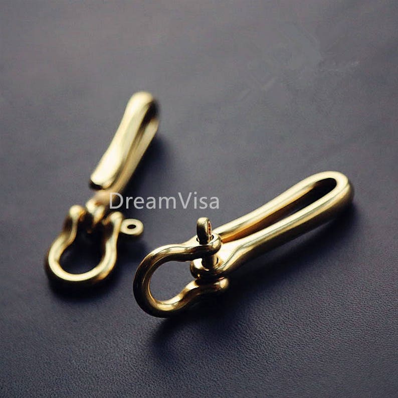 Op de afbeelding: Twee goudkleurige metalen sleutelhangers met een haak en een draaibare sluiting. De sleutelhangers zijn gemaakt van massief messing en hebben een gepolijste afwerking.