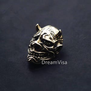 Könnte beinhalten: Ein goldfarbener Metallanhänger in Form einer japanischen Oni-Maske mit Hörnern und einem grimmigen Ausdruck.