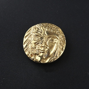 Könnte beinhalten: Ein runder, goldfarbener Knopf mit einem detaillierten Relief eines Gesichts. Das Gesicht hat einen Bart und komplizierte Haare, mit einem leicht strengen Ausdruck. Der Knopf ist vor einem schwarzen Hintergrund platziert.