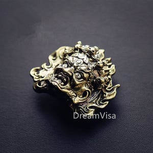 Puede incluir: Un colgante de metal dorado que presenta una representación detallada de una criatura mítica con múltiples ojos y una expresión feroz. La criatura está rodeada de llamas intrincadas y diseños florales.