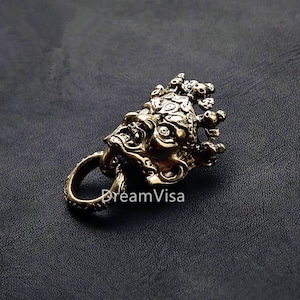 Könnte beinhalten: Ein goldfarbener Schlüsselanhänger aus Metall mit einem detaillierten, kunstvollen Design eines Dämonengesichts mit einer Krone. Der Anhänger hat einen Ring zum Anbringen an einem Schlüsselbund.