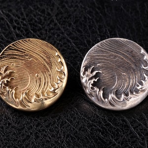 Puede incluir: Dos botones redondos de metal con un diseño de ola. Un botón es dorado y el otro es plateado.