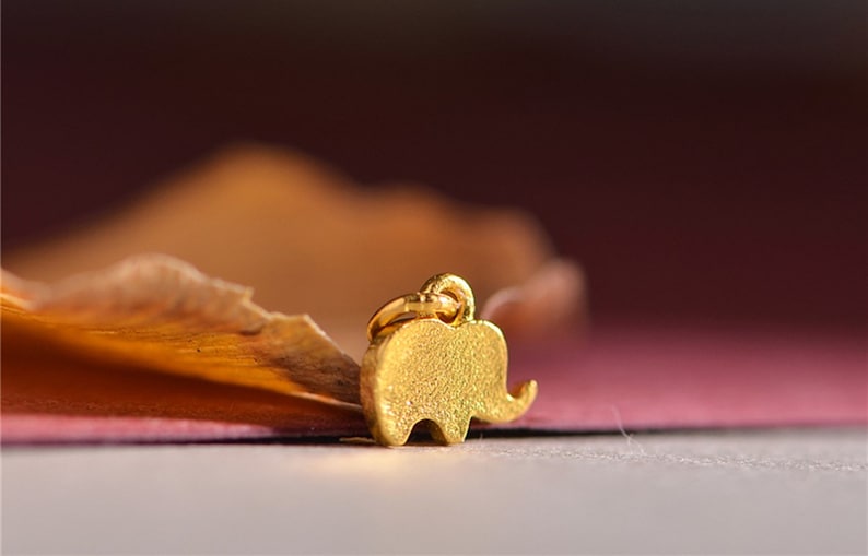 May include: A small, gold-colored elephant charm with a textured surface. The charm has a loop at the top for attaching to a necklace or bracelet. The background is a blurred mix of brown, orange, and pink.
