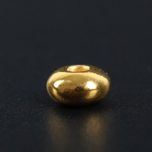 May include: A single, shiny, gold-colored bead with a rounded, donut-like shape. The bead has a smooth, reflective surface and a small, central hole. The background is a dark, solid color.