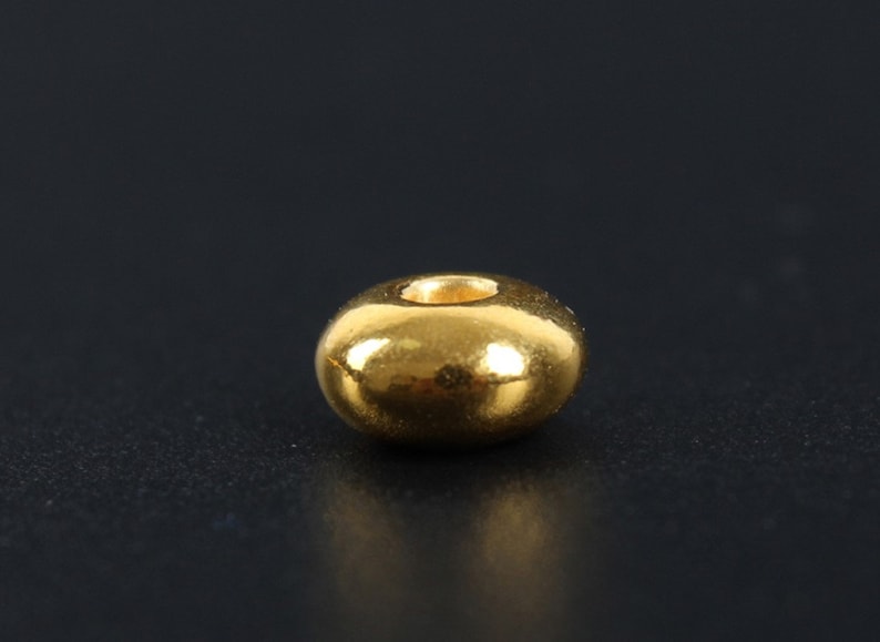 May include: A single, shiny, gold-colored bead with a rounded, donut-like shape. The bead has a smooth, reflective surface and a small, central hole. The background is a dark, solid color.