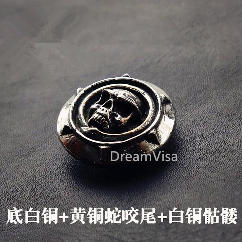 May include: A silver skull pendant with a detailed design. The skull is surrounded by a circle with a starburst pattern. The pendant is on a dark gray background.