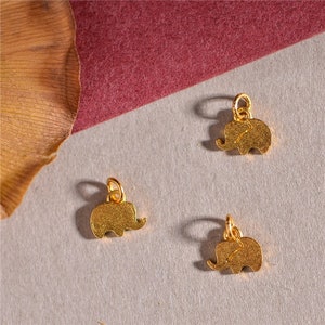 May include: Three small, gold-colored elephant charms with a textured surface. Each charm has a loop at the top for attaching to a necklace or bracelet. The charms are displayed on a gray surface with a maroon background.