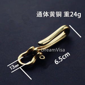 Op de afbeelding: Een gouden metalen haak met een lus aan het ene uiteinde en een haak aan het andere uiteinde. De haak is 6,5 cm lang en 1,3 cm breed. Hij weegt 24 gram.