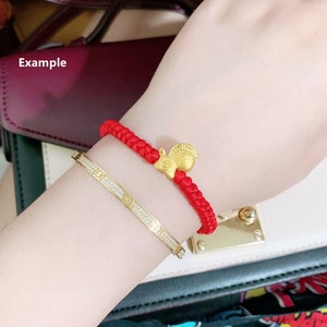 May include: A red string bracelet with a gold squirrel charm. The bracelet is worn on a person's wrist, along with a gold bangle bracelet with diamonds.