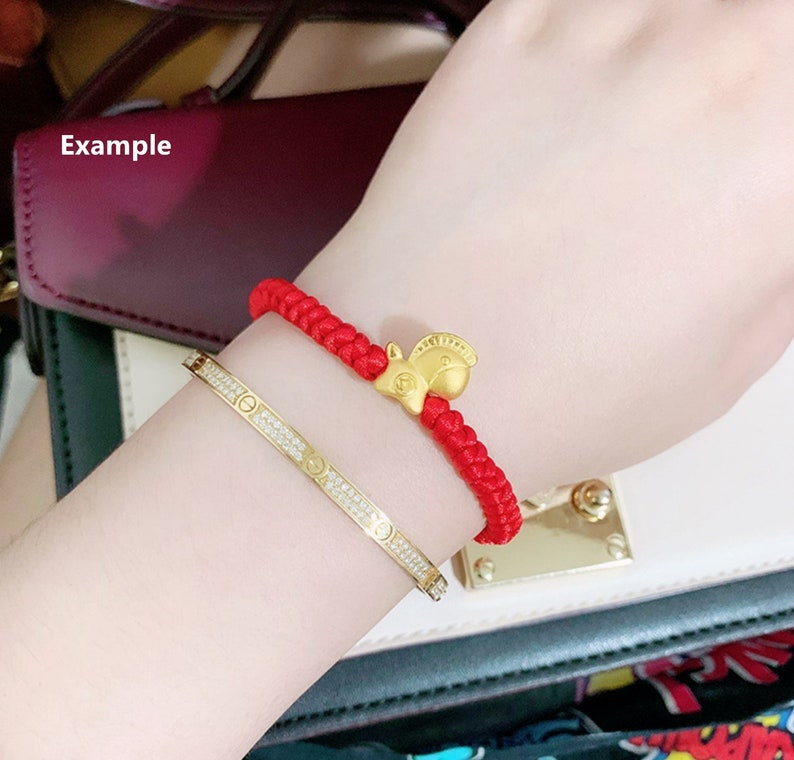 May include: A red string bracelet with a gold squirrel charm. The bracelet is worn on a person's wrist, along with a gold bangle bracelet with diamonds.