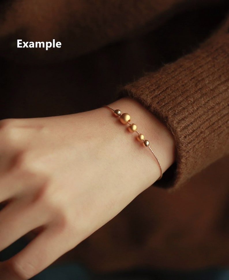 K&ouml;nnte beinhalten: Ein zartes goldenes Armband mit einer Kette und einer Reihe von abgestuften goldenen Perlen.