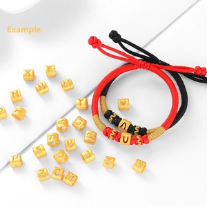 May include: Two red and black string bracelets with gold letter beads spelling "AU" and a heart charm.  A scattering of gold letter beads in the background.