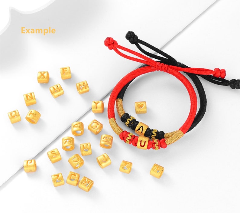 May include: Two red and black string bracelets with gold letter beads spelling "AU" and a heart charm.  A scattering of gold letter beads in the background.