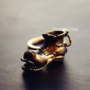 Könnte beinhalten: Ein goldfarbener Schlüsselanhänger in Form eines Stiefels. Der Stiefel weist aufwendige Details auf, darunter Schnürsenkel und eine Schlaufe zur Befestigung. Der Schlüsselanhänger besteht aus glänzendem Metall und ist vor einem dunklen Hintergrund platziert.