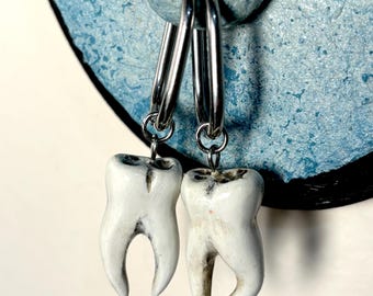 Puede incluir: Un par de pendientes de aro plateados con dijes de dientes blancos que cuelgan de ellos. Los pendientes están colgados sobre un fondo azul y negro.