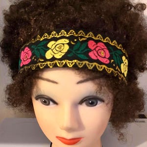 以下が含まれることがあります： 茶色の巻き毛と黒いヘアバンドをつけたマネキンの頭。ヘアバンドには、黄色とピンクのバラ、緑の葉、金のトリムが刺繍されています。マネキンは、明るい肌色、赤い口紅、濃いアイメイクをしています。