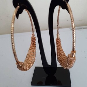 May include: A pair of gold hoop earrings with a woven design. The earrings are displayed on a black stand.