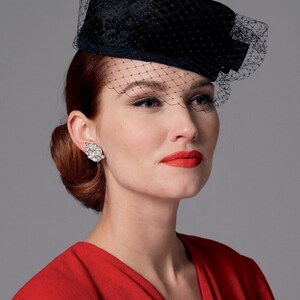 May include: A black hat with a veil, a vintage-inspired accessory. The hat is worn by a person with red hair and red lipstick. The hat is a rectangular shape with a net veil. The person is wearing a red dress.