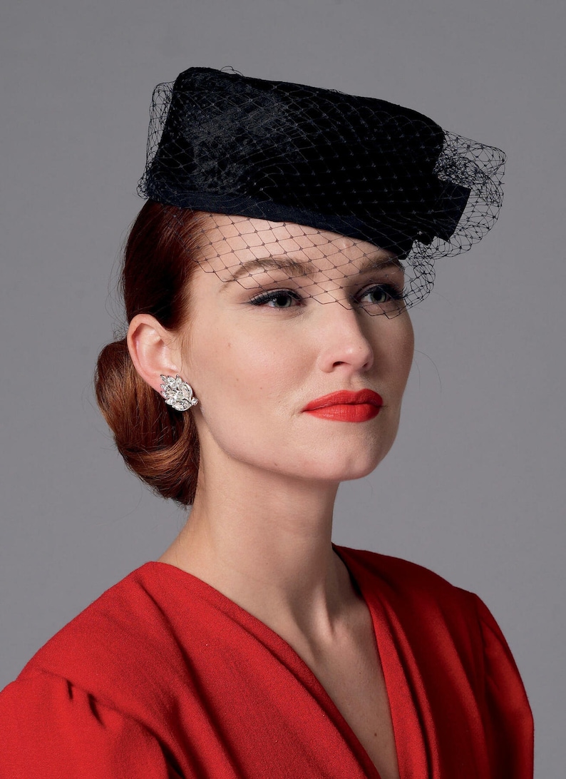 May include: A black hat with a veil, a vintage-inspired accessory. The hat is worn by a person with red hair and red lipstick. The hat is a rectangular shape with a net veil. The person is wearing a red dress.