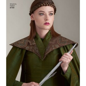 May include: A woman wearing a green dress with a brown patterned collar and a silver headband. She is holding a silver arrow in her right hand.