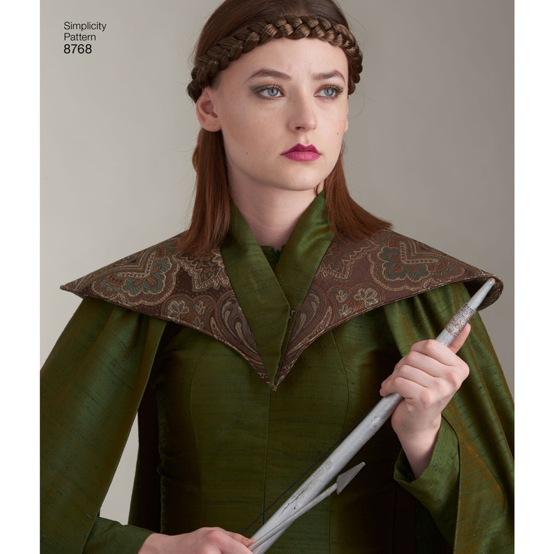 May include: A woman wearing a green dress with a brown patterned collar and a silver headband. She is holding a silver arrow in her right hand.