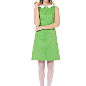 May include: A sleeveless green dress with a white Peter Pan collar and white buttons. The dress has a fitted bodice and a slightly flared skirt. The model is wearing white tights and blue shoes.