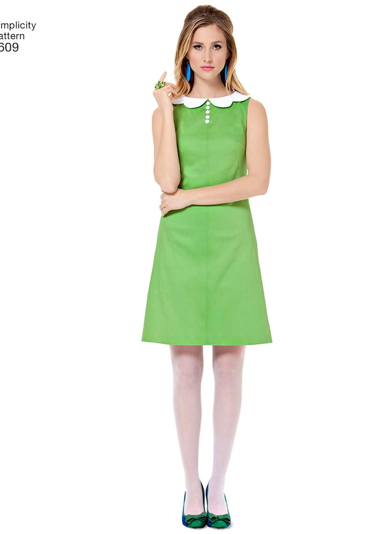 May include: A sleeveless green dress with a white Peter Pan collar and white buttons. The dress has a fitted bodice and a slightly flared skirt. The model is wearing white tights and blue shoes.