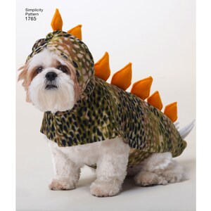 May include: A white dog wearing a green and brown dinosaur costume with orange spikes. The costume has a hood and a tail.