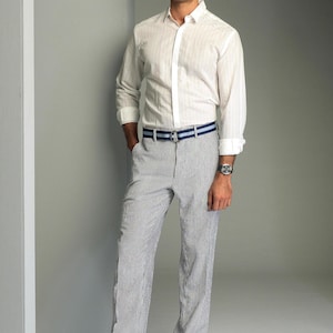 May include: A white button-down shirt with a striped pattern and a blue and white striped belt. The man is wearing white shoes.