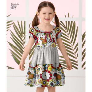 Pu&ograve; includere: Una bambina che indossa un vestito floreale bianco, rosso e giallo con un fiocco rosso al collo. Il vestito ha un motivo a quadri bianchi e neri al centro. Simplicity Pattern 2377 &egrave; stampato nell'angolo in alto a sinistra dell'immagine.