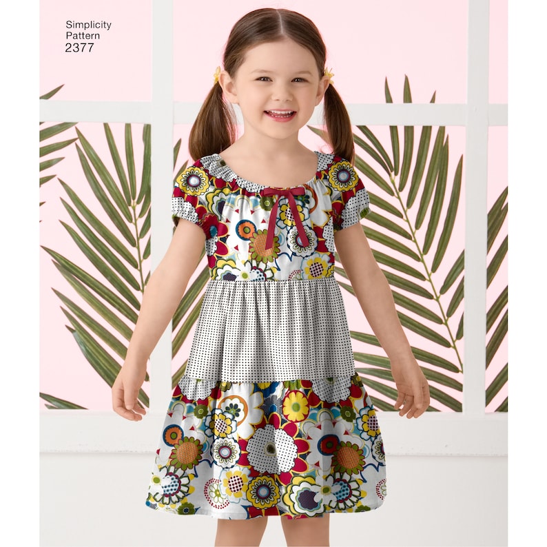 Pu&ograve; includere: Una bambina che indossa un vestito floreale bianco, rosso e giallo con un fiocco rosso al collo. Il vestito ha un motivo a quadri bianchi e neri al centro. Simplicity Pattern 2377 &egrave; stampato nell'angolo in alto a sinistra dell'immagine.