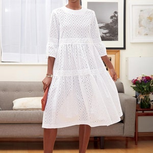 May include: A white eyelet dress with a tiered skirt and three-quarter sleeves. The dress features a round neckline and is paired with patterned heels and a coral clutch. The dress is knee-length.