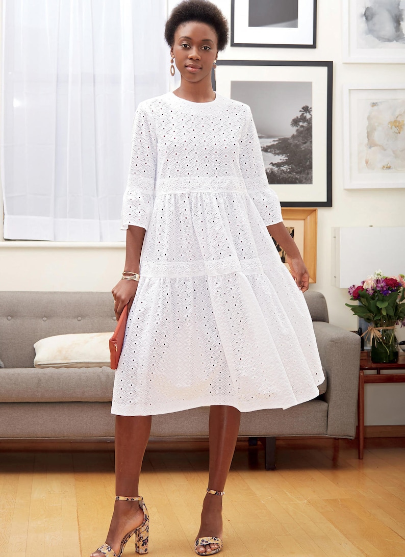May include: A white eyelet dress with a tiered skirt and three-quarter sleeves. The dress features a round neckline and is paired with patterned heels and a coral clutch. The dress is knee-length.