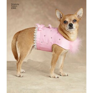 May include: A small dog wearing a pink dog dress with a white lace trim and small bows. The dress is adorned with rhinestones and a fluffy pink feather collar. The dog is standing on a neutral-colored surface.