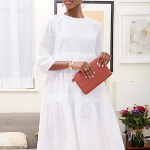 May include: A white eyelet midi dress with three-quarter sleeves and a tiered skirt. The dress is paired with a rust-colored leather clutch. The model is wearing gold jewelry, including earrings and a bracelet. The background includes framed artwork and a sofa.