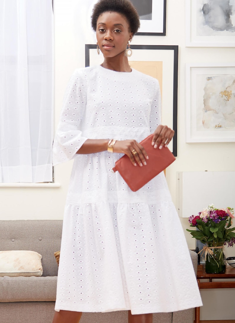 May include: A white eyelet midi dress with three-quarter sleeves and a tiered skirt. The dress is paired with a rust-colored leather clutch. The model is wearing gold jewelry, including earrings and a bracelet. The background includes framed artwork and a sofa.