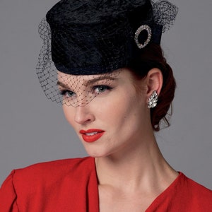 May include: A black velvet pillbox hat with a delicate net veil. The hat features a sparkling, circular rhinestone embellishment. The woman in the image is wearing a red dress and a pair of sparkling earrings.