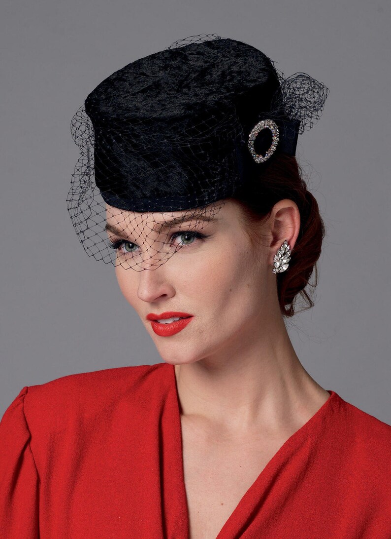 May include: A black velvet pillbox hat with a delicate net veil. The hat features a sparkling, circular rhinestone embellishment. The woman in the image is wearing a red dress and a pair of sparkling earrings.