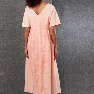 May include: A peach-colored midi dress with a V-neck and short sleeves. The dress features a gathered waist and vertical seams down the front. The fabric has a subtle, mottled pattern. The model is wearing brown sandals.