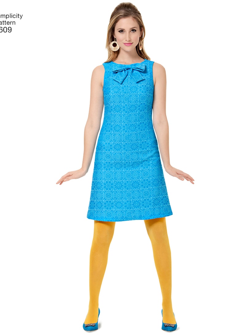 May include: A sleeveless blue dress with a white pattern and a bow at the neckline. The dress is fitted at the waist and flares out slightly at the hem. The model is wearing yellow tights and blue shoes.