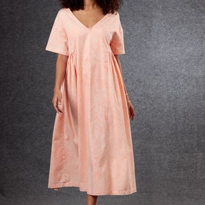 May include: A peach-colored midi dress with a V-neck and short sleeves. The dress features a gathered waist and vertical seams down the front. The model is wearing the dress with sandals.