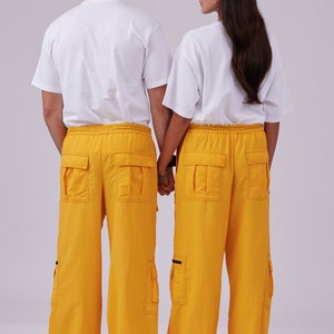 May include: Two people wearing white t-shirts and matching yellow cargo pants with black accents. They are standing back to back with their hands clasped together.
