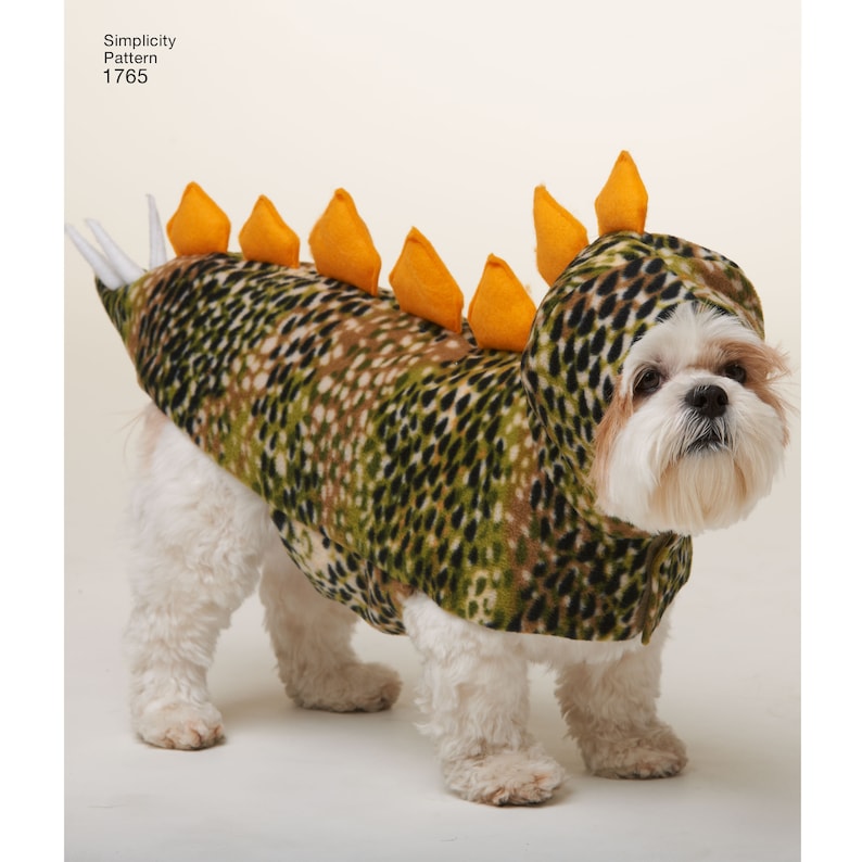 May include: A white dog wearing a green and brown camouflage dinosaur costume with orange spikes on the back.