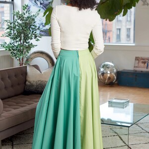 May include: A woman wearing a white long-sleeved top and a green and light green maxi skirt. The skirt is split down the middle, with one side being a darker green and the other side being a lighter green.