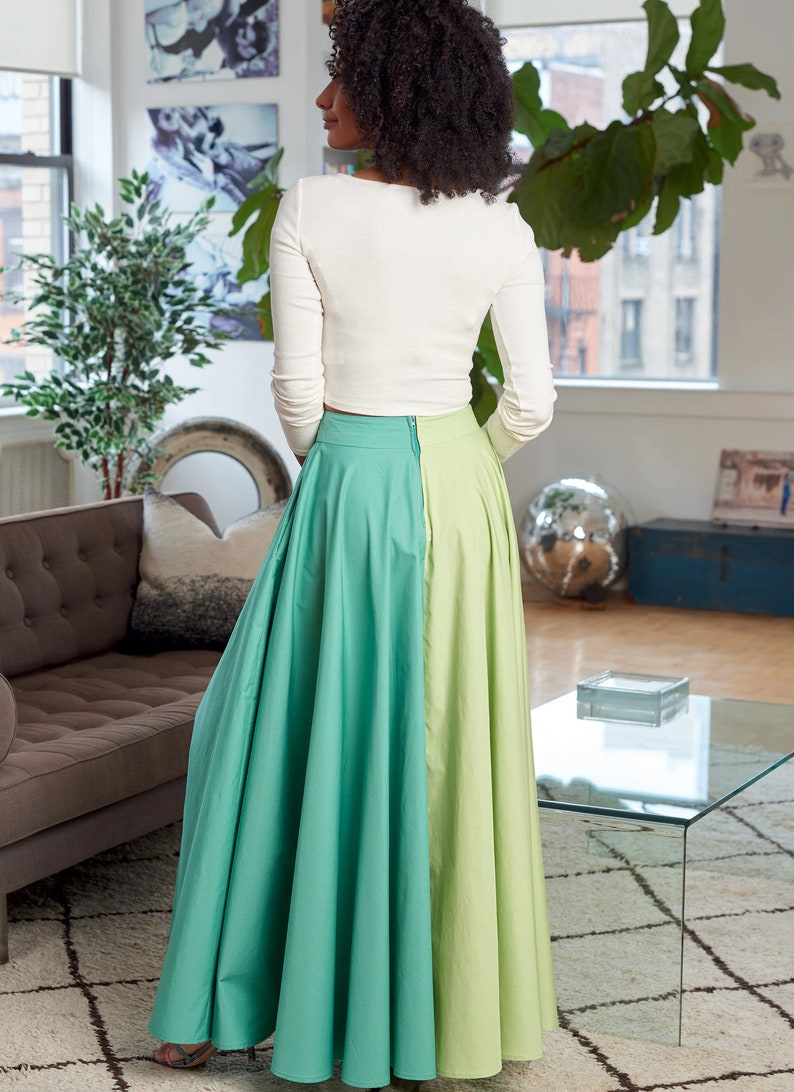 May include: A woman wearing a white long-sleeved top and a green and light green maxi skirt. The skirt is split down the middle, with one side being a darker green and the other side being a lighter green.