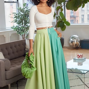 May include: A woman wearing a white long-sleeved crop top with a tie at the waist and a green and blue maxi skirt. She is holding a green handbag.